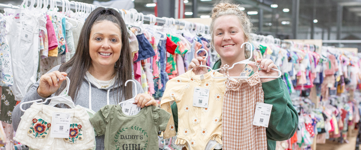 Two ladies smile as the hold up hangers with clothing on them. They are both standing next to a rack full of clothing for sale.